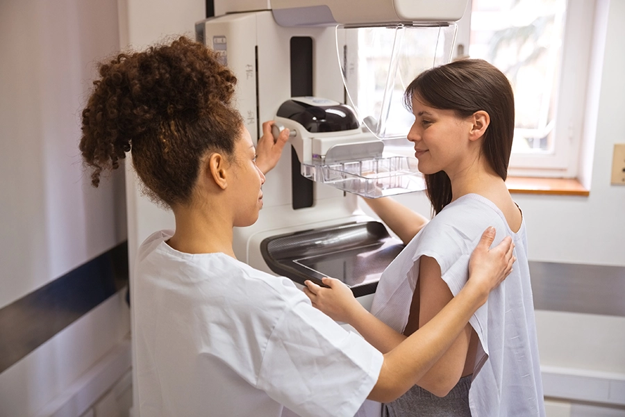 Mammography technologist helps a smiling woman in a hospital gown position for a mammogram.